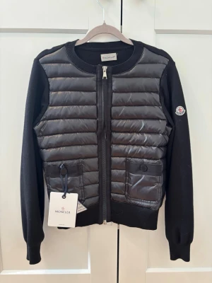 Moncler Cardigan - Knappt använd svart Moncler Cardigan, köpt second hand men i toppenskick. En enda defekt är att lappen är trasig, visas i bild 7. Moncler storlek 2. QR kod samt äkthetskod visas i bild 3. Perfekt som trendig höstjacka! 🍂