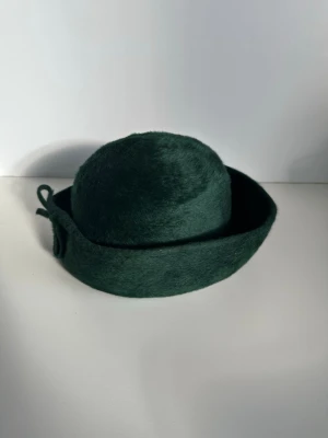 Adela Isakson - grön hatt i filt med rosett - storlek S /53cm - Stilren grön hatt i filt från legendariska svenska hattmakerskan / hattkonstnären Adela Isakson från Borås. Hatten har en rundad kulle och ett brett brätte som är uppvikt runt om. En liten stilfull rosett pryder baksidan av hatten. Len borstad mörkgrön filt, för mjuk känsla. Passar huvudomkrets på 53cm, motsvarande ca storlek S/M. Klassisk och tidlös design som passar för olika tillfällen. Ett stycke designhistoria i hattform. Mycket gott skick, som ny!