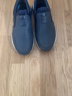 Lv skor - Snygga mörkblå slip-on sneakers med vit sula och diskret logga på plösen. Skorna har en minimalistisk design utan snörning och är tillverkade i ett slätt material som ger en clean look. Perfekta för dig som gillar enkel och stilren streetwear.LV