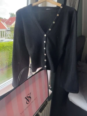 Svart kofta med pärlemorknappar - Stilren svart kofta från Gina Tricot med v-ringning och pärlemorskimrande knappar längs hela framsidan. Modellen har långa ärmar och ribbad midja som ger en snygg siluett. Perfekt att matcha med jeans eller kjol för en trendig look.