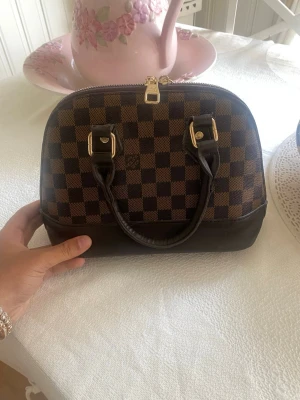  Alma handväska - Snygg handväska i klassiskt brunt Damier-mönster. Väskan har rundad form, två mörkbruna handtag och guldfärgade detaljer. Tillverkad i canvas och läder.