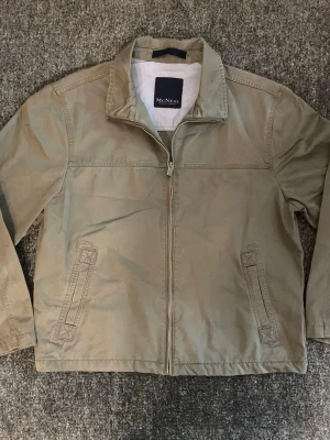 Beige vindjacka från McNeal - Stilren beige vindjacka från McNeal med dragkedja framtill och två snedställda fickor. Jackan har klassisk krage och är tillverkad i ett lätt material som passar perfekt för blåsiga dagar. Enkel och tidlös design som funkar till det mesta.