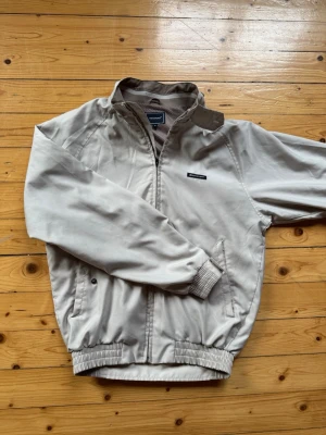 Beige vindjacka från Rockport - Snygg beige vindjacka från Rockport med dragkedja, ståkrage och ribbade muddar. Jackan har två sidofickor och meshfoder på insidan för extra komfort. Perfekt för blåsiga dagar och enkel att matcha med olika outfits.