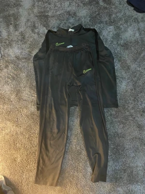 Nike svart träningsset med ziptröja - Säljer ett svart träningsset från Nike med långärmad ziptröja och matchande byxor. Båda plaggen har neongula Nike-loggor och är tillverkade i ett stretchigt, snabbtorkande material som passar perfekt till träning eller uppvärmning.