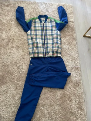 Rutigt träningsset med blå byxor - Snyggt träningsset från Lacoste med jacka i rutigt mönster i blått, grönt, vitt och rött samt matchande blå byxor. Jackan har dragkedja och muddar vid ärmslut, medan byxorna har resår i midjan och detaljer i samma rutiga tyg. Perfekt för sport eller chill.