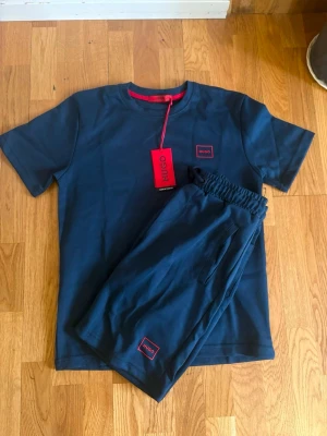 Mörkblått set från Hugo - Snyggt mörkblått set från Hugo med t-shirt och shorts. Båda plaggen har diskreta röda Hugo-loggor. T-shirten har rund hals och korta ärmar, shortsen har resår i midjan och sidofickor. Materialet ser ut att vara mjukt och bekvämt, perfekt för chill dagar.