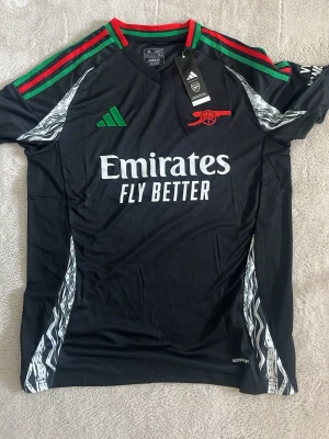 Svart Arsenal matchtröja Adidas - Snygg svart Arsenal matchtröja från Adidas med gröna och röda detaljer på axlarna. Tryck med Emirates Fly Better på bröstet och klubbmärke i rött. Tröjan har korta ärmar och vita grafiska mönster på sidorna. Perfekt för fotbollsträning eller match.