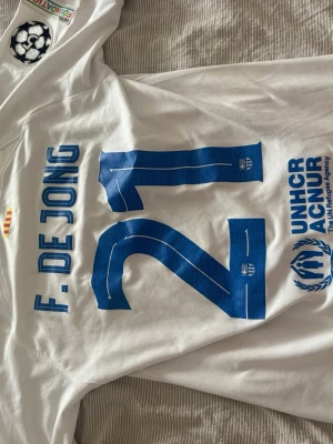 Fotbollströja - Säljer en vit FC Barcelona matchtröja med blå detaljer, tryck 'F. DE JONG 21' på ryggen och officiella loggor som Spotify och UNHCR. Tröjan har klubbmärke på bröstet och Champions League-märke på ärmen. Perfekt för fotbollsfans!