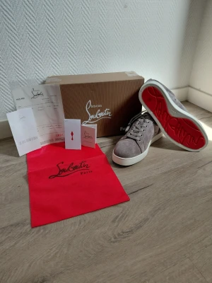 Christian Louboutin - Snygga gråa sneakers från Christian Louboutin i mjuk mocka med klassisk röd sula och vita kanter. Skorna har lågt skaft, matchande gråa skosnören och diskreta sömmar för en clean look. Perfekta för dig som vill ha något exklusivt och stilrent.