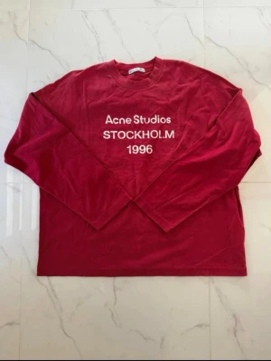 Röd långärmad topp Acne Studios - Cool röd långärmad topp från Acne Studios med tryck 'Acne Studios STOCKHOLM 1996' på bröstet. Klassisk rund halsringning och relaxed fit. Perfekt för dig som gillar streetwear och vill ha något stilrent men ändå statement.