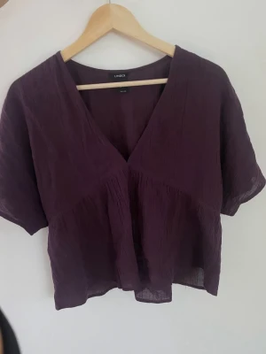 Plommonlila blus med v-ringning Lindex - Plommonlila blus från Lindex med djup v-ringning och korta ärmar. Tillverkad i ett tunt, lätt och något skrynkligt tyg som ger en avslappnad känsla. Perfekt att styla med både jeans och kjol för en trendig look.