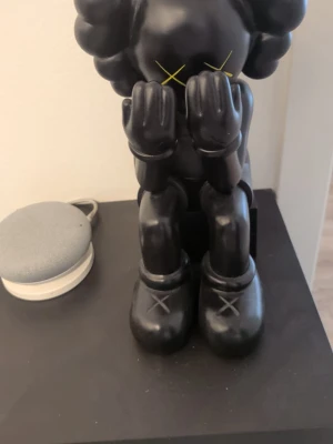 Svart KAWS-figur - Säljer en svart sittande konstfigur av KAWS, med gula kors på ögonen och händerna för ansiktet. Figuren har rundade former och stora skor med X på tårna. Tillverkad i hård plast, perfekt som dekoration i hemmet.