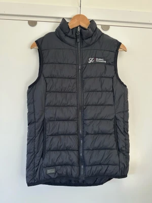 South West - Svart vadderad väst med hög krage - storlek M - Stilren svart vadderad väst från South West Everywear i storlek M. Tidlös modell med dragkedja framtill och hög krage. Liten logotyp nedtill på vänster sida. Västen är designad för att ge extra värme och komfort utan att vara för skrymmande. Len vattenavvisande nylon. Fint skick!!