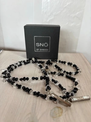 Svart och transparent pärlhalsband SNÖ - Säljer ett stilrent halsband från SNÖ of Sweden med små svarta och transparenta pärlor. Halsbandet har ett silverfärgat hänge med märkets logga. Perfekt accessoar för att lyfta din outfit.
