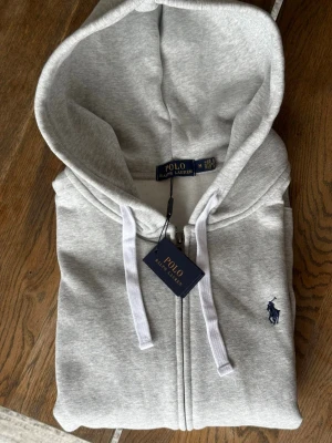  Polo Ralph Lauren Hoodie - Snygg grå hoodie från Polo Ralph Lauren med dragkedja, vita snören i huvan och klassisk mörkblå logga broderad på bröstet. Tillverkad i mjuk bomullsblandning och har känguruficka framtill. Perfekt för en avslappnad och stilren look.