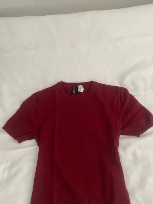 Vinröd stickad topp med kort ärm - Säljer en vinröd stickad topp med korta ärmar och rund hals. Toppen har ribbade muddar vid ärmslut och nederkant för en snygg passform. Perfekt att styla med jeans eller kjol för en trendig look💕