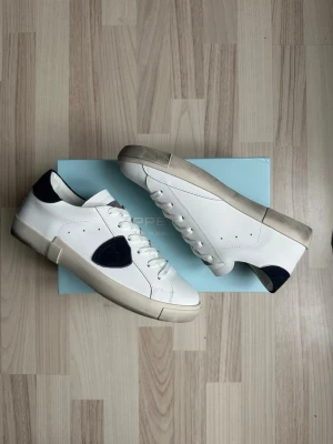 Vita sneakers från Philippe Model - Snygga vita sneakers från Philippe Model med mörkblå detaljer på häl och sidan. Skorna har en klassisk låg siluett, vita snören och en robust sula i gummi. Tillverkade i slätt läder med diskreta hål för ventilation. Perfekta för en clean och stilren look.