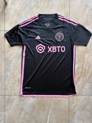 Svart Inter Miami matchtröja Adidas - Säljer en svart Inter Miami matchtröja från Adidas med rosa detaljer och logga på bröstet. Tröjan har korta ärmar, rund hals och rosa ränder på axlarna. Tillverkad i lätt och ventilerande material, perfekt för fotboll eller träning.
