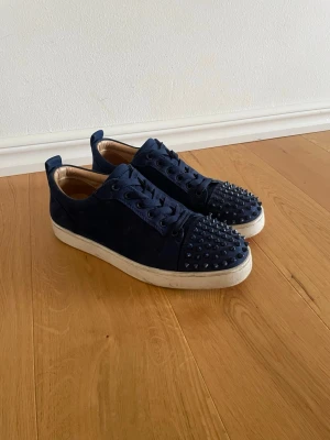Louboutin - Snygga mörkblå sneakers i mocka med coola mörkblå nitar på tån och matchande mörkblå skosnören. Skorna har vit sula och en låg profil som ger en modern look. Perfekta för dig som vill sticka ut med din stil. Storlek 43 men passar även 42. Priset är inte hugget i sten! 