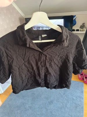 Svart croppad skjorttopp - Säljer en svart croppad skjorttopp med korta ärmar och krage. Toppen har knappar framtill och är tillverkad i ett mjukt bomullsliknande material. Perfekt för dig som gillar en avslappnad men ändå trendig stil.