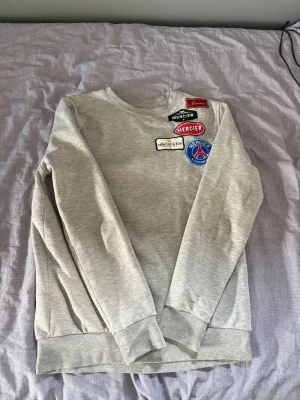 Mercier Tracksuit - Säljer en grå sweatshirt med flera coola patches på bröstet i rött, blått, vitt och svart. Tröjan har rund halsringning, långa ärmar och ribbade muddar vid ärmslut och nederkant. Perfekt för dig som gillar streetwear och unika detaljer.