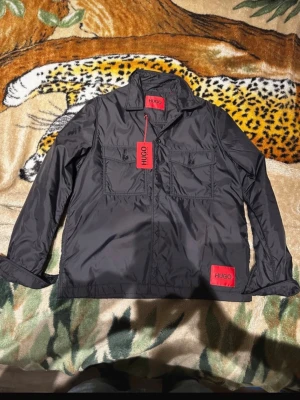 Svart overshirt från HUGO - Stilren svart overshirt från HUGO med två stora bröstfickor och röda HUGO-detaljer. Overshirten har klassisk krage, raka ärmar och är tillverkad i ett glansigt, lätt material som ger en modern look. Perfekt för lager-på-lager och streetwear.