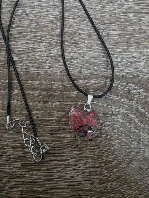 Halsband med hjärthänge och blommor - Unikt halsband med svart snöre och ett hjärtformat hänge i transparent glas. Hänget innehåller små röda blommor med fjäril och svarta detaljer som ger en cool och romantisk vibe. Perfekt accessoar för dig som vill sticka ut med något personligt och kreativt.