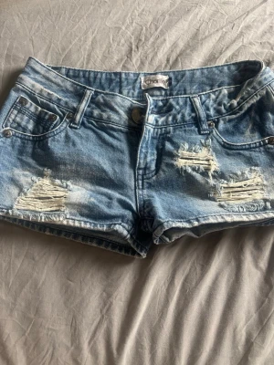 Blå slitna jeansshorts - Säljer ett par blå jeansshorts med slitna detaljer. Shortsen har klassisk femficksdesign, dragkedja och knapp framtill samt coola bakfickor med dragkedjor. Perfekta för sommaren och ger en avslappnad streetstyle. Passar någon som bär xs-s, står s på lappen