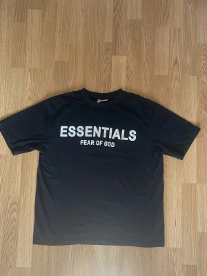 Svart Essentials Fear of God t-shirt - Svart t-shirt från Essentials Fear of God med vit text på bröstet. Klassisk passform med rund hals och korta ärmar. Tillverkad i mjuk bomull som känns skön mot huden. Perfekt för en stilren och avslappnad look.
