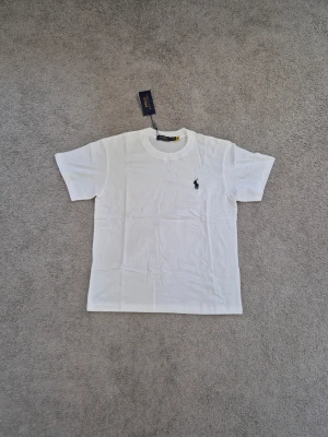 Polo Ralph Lauren - Klassisk vit t-shirt från Polo Ralph Lauren med den ikoniska svarta loggan broderad på bröstet. T-shirten har rund halsringning och korta ärmar. Tillverkad i mjuk bomull för en skön och avslappnad stil.