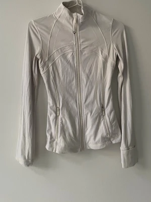 Vit definejaket från Lululemon - Snygg och figurnära vit zip-jacka från Lululemon med hög krage och långa ärmar. Jackan har två fickor med dragkedja och dekorativa sömmar som ger en sportig look. Perfekt för träning eller som ett extra lager. Materialet är stretchigt och mjukt.