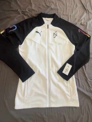 Puma träningströjar Neymar JR limited Edition - Säljer en snygg träningsjacka från Puma i vitt och svart med hel dragkedja. Jackan har färgglada detaljer på ärmarna och Puma-logga på bröstet. Tillverkad i ett mjukt och stretchigt material som passar perfekt till träning eller sport.