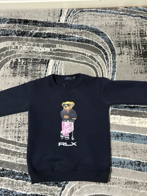 Mörkblå sweatshirt med björntryck RLX - Säljer en mörkblå sweatshirt med ett stort tryck av en björn i streetwear-stil på bröstet och RLX-logga under. Tröjan har rund halsringning och långa ärmar. Perfekt för dig som gillar coola och unika prints.