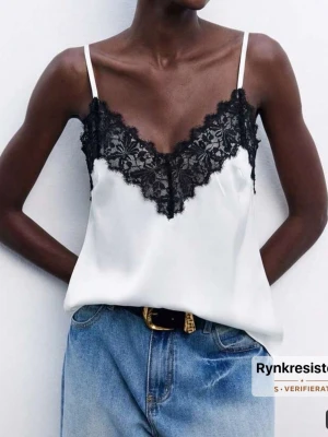 Vit linnetopp med svart spets - Snygg vit linnetopp med tunna axelband och dekorativ svart spets längs v-ringningen. Toppen har en avslappnad passform och är perfekt att styla med jeans eller kjol för en trendig look.