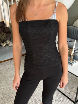 Svart spetskorsett med axelband - Snygg svart korsettopp med spetsdetaljer och smala axelband. Toppen har en figurnära passform och är perfekt för att lyfta vilken outfit som helst. Spetsen ger en elegant touch och den raka skärningen framtill gör den extra stilren.