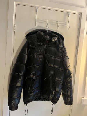 Svart dunjacka från Moncler med huva - Snygg svart dunjacka från Moncler i glansigt material. Jackan har avtagbar huva, dragkedjor på fickorna och justerbar nederkant med snören. Perfekt för kalla vinterdagar och riktigt trendig look.