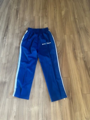 Blåa Palm Angels mjukisbyxor - Säljer ett par blåa mjukisbyxor från Palm Angels med vita sidoränder och logga på vänster ben. Byxorna har resår i midjan och raka ben. Perfekta för en avslappnad och trendig streetwear-look. storlek S passar 170-180 pris går att diskutera 