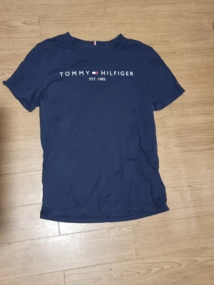 Marinblå Tommy Hilfiger t-shirt - INSTABOX!!! Snygg marinblå t-shirt från Tommy Hilfiger med klassisk logga och texten 'EST. 1985' på bröstet. T-shirten har rund hals och korta ärmar, perfekt för en avslappnad och stilren look.