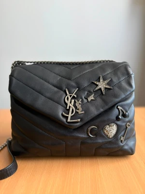 Svart YSL handväska med kedja - Snygg svart handväska från YSL i mjukt läder med quiltad design och silverfärgade detaljer. Väskan har dekorativa pins i form av stjärna, hjärta, måne och nyckel samt en kedjerem. Perfekt accessoar för att lyfta din outfit.