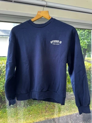 Mörkblå sweatshirt Studio 10 - Mörkblå sweatshirt med rund hals och vitt tryck på bröstet. Tröjan har ribbade muddar vid ärmslut och nederkant samt en avslappnad passform. Tillverkad i mjukt material, perfekt för en chill och sportig stil. Storlek S, men den är oversized. 