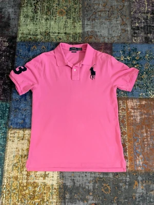 Rosa pikétröja från Ralph Lauren - Säljer en klassisk rosa pikétröja från Ralph Lauren med korta ärmar och krage. Tröjan har en stor mörkblå logga på bröstet och siffran 3 på ena ärmen. Perfekt för dig som vill ha en sportig och stilren look. OBS: den passar som en M
