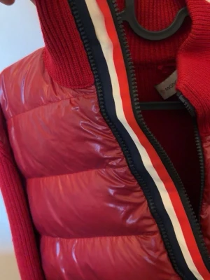 Röd dunväst från Moncler - Snygg röd dunväst från Moncler med stickade detaljer och Moncler-logga på ärmen. Västen har en dragkedja med röd, vit och marinblå rand längs kanten. Perfekt för dig som vill ha en sportig och trendig look.