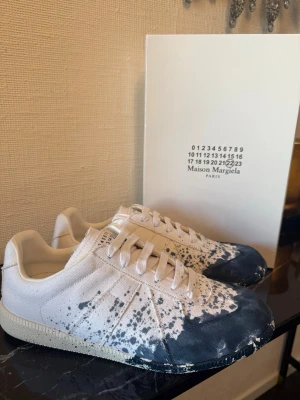 Maison Margiela med blå stänk - Maison Margiela sneakers i vitt tyg med unika blå färgstänk över hela skon. Klassisk låg modell med snörning och vit sula. Finns kvitto och box.