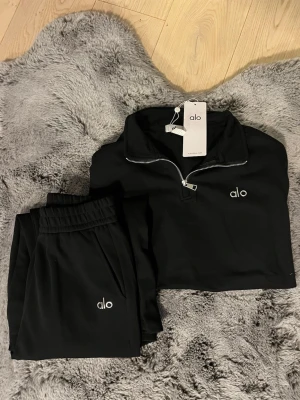 Svart tracksuit från Alo - Svarta mjukisbyxor från Alo med avslappnad passform och elastisk midja. Byxorna har en silvrig Alo-logga framtill och är gjorda i ett mjukt och bekvämt material. Perfekta för chill eller träning. Den passar storlek M för en mer ”perfekt” passform och S för en mer loose passform