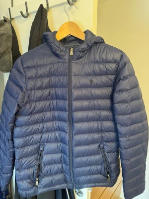 Mörkblå pufferjacka från Ralph Lauren - Snygg mörkblå pufferjacka med huva och dragkedja framtill. Den är från Ralph Lauren och är perfekt till vintern! Jag vet faktiskt inte vilken storlek men modellen på bilden är 181 cm lång så är man runt där så passar den! Tror den är XL för barn.