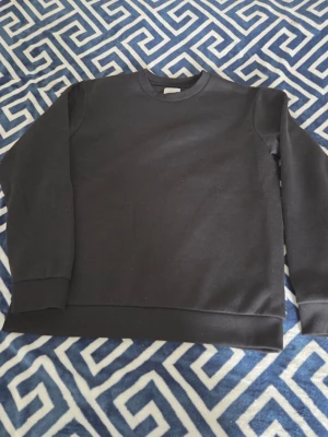 Svart stickad sweatshirt - En enkel och stilren svart stickad sweatshirt med rund hals och långa ärmar. Perfekt basplagg som passar till det mesta och är lätt att matcha med andra kläder. Ribbstickade muddar vid ärmslut och nederkant.