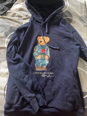 Marinblå hoodie med björntryck (Ralph Lauren) - Snygg marinblå hoodie från Ralph Lauren med ikoniskt björntryck på bröstet. Hoodien har dragsko i huvan och ribbade muddar. Perfekt för en avslappnad och trendig stil. Materialet känns mjukt och bekvämt mot huden.