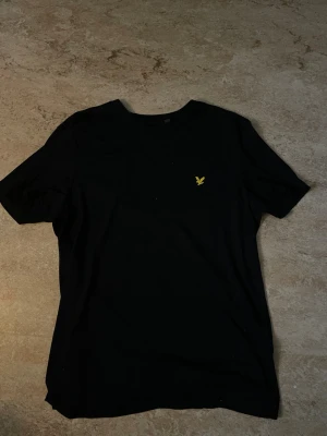 Svart t-shirt med gul broderad logga - Snygg svart t-shirt med en liten gul broderad logga på bröstet. Klassisk passform och rund halsringning. Perfekt basplagg som funkar till allt och är enkel att styla. Tillverkad i mjuk bomull för skön känsla hela dagen. Lyle and scott, saknar storlek anvisning men skulle säga att den passar L/M