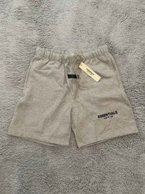 Gråa Essentials shorts med snörning - Snygga gråa shorts från Essentials med elastisk midja och snörning framtill. Shortsen har en enkel design med logga på vänster ben och är tillverkade i mjuk bomull. Perfekta för chill dagar eller när du vill ha en clean look.