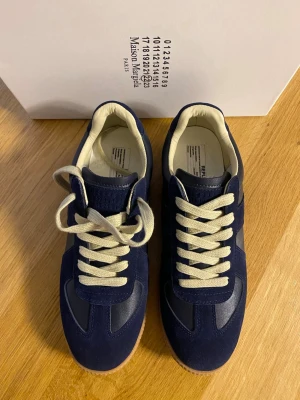 Mörkblå sneakers Maison Margiela - Snygga mörkblå sneakers från Maison Margiela med ovandel i mocka och läder, vita snören och beige innersula. Klassisk låg modell med gummisula och diskreta detaljer. Perfekta för dig som vill ha en stilren och trendig look.